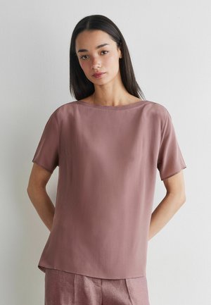 Ung kvinde med langt mørkt hår iført en lilla kortærmet bluse og matchende bukser mod en ensfarvet hvid baggrund.