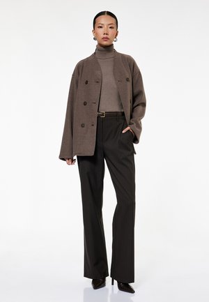 Donna in piedi che indossa una giacca marrone con bottoni, dolcevita marrone, pantaloni ampi marrone scuro e tacchi neri a punta su sfondo bianco.