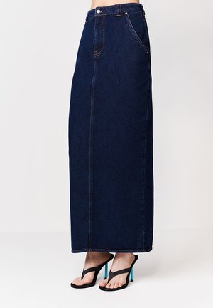Tmavomodrá dlhá sukňa z džínsoviny s vysokým pásom a predným švom, kombinovaná s čiernymi vysokými sandálmi na podpätku s jasne modrými ihličkovými podpätkami.