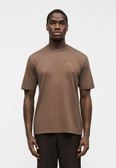 Homme portant un t-shirt à manches courtes marron avec un petit logo blanc "NN. 07" sur la poitrine, debout les bras détendus devant un fond blanc uni.