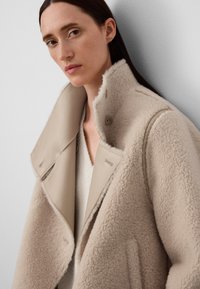 Beżowy płaszcz z owczej wełny z skórzanym kołnierzem, z podwójnym zapięciem i widocznymi guzikami. Miękka tekstura i minimalne detale szycia.