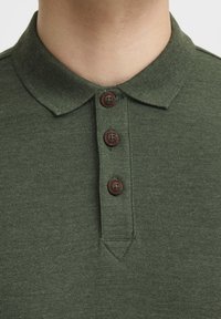 Grøn struktureret polo-shirt med krave, tre brune knapper og en stolpe. Stoffet ser blødt ud og er jævnt vævet.