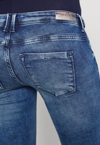 Jeans de denim azul con un corte ajustado, bolsillos traseros y una etiqueta de cuero de la marca. Tela texturizada con un ligero desgaste y efectos de arrugado.
