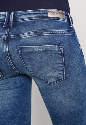 Jeans de denim azul con un corte ajustado, bolsillos traseros y una etiqueta de cuero de la marca. Tela texturizada con un ligero desgaste y efectos de arrugado.