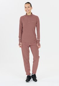 Vrouw in een oud-roze lange mouw sporttop en bijpassende broek, staand en lachend, met zwart-witte hardloopschoenen op een witte achtergrond.