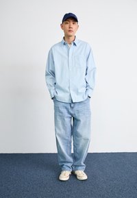 Lichtblauwe gestreepte button-up shirt met lange mouwen, gecombineerd met loszittende lichtblauwe jeans en witte sneakers. Donkerblauwe pet gedragen.