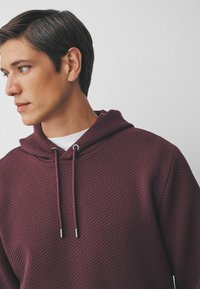 Burgunderfarbener Hoodie mit einem strukturierten Rautenmuster, ausgestattet mit einem Zugband mit metallischen Spitzen und einem weißen Shirt darunter.