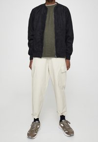 Veste aviateur en daim noir, chemise texturée vert olive, pantalon cargo crème, et baskets marron avec des accents. Coupe structurée.