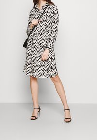 Black and white chevron-patterned shirt dress with long sleeves, cinched waist, and button-up collar, paired with black strappy heels.
