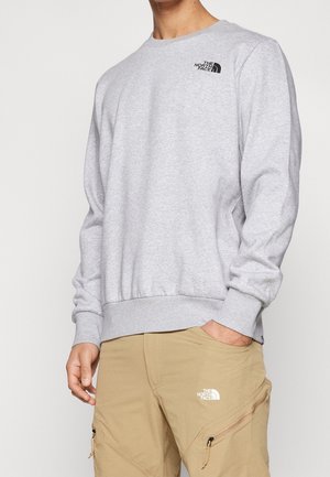 Osoba ubrana w jasnoszarą bluzę z długim rękawem oraz jasnobrązowe spodnie typu cargo, na obu widoczne logo The North Face na piersi i udzie.