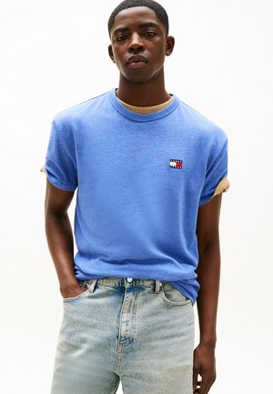 Blue cotton t-shirt with rolled sleeves, featuring a small embroidered logo in red, white, and navy on the chest. Worn with light blue jeans.