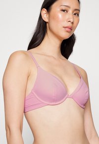 Sutiã acolchoado rosa claro com uma textura suave, alças finas e ajustáveis, e um formato triangular, apresentando um fecho simples nas costas.