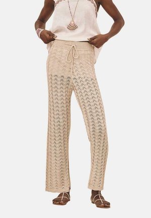 Pantalones de lounge de punto beige con cintura ajustable, que presentan un patrón abierto similar al encaje y un diseño de pierna ancha, estilizados con sandalias.
