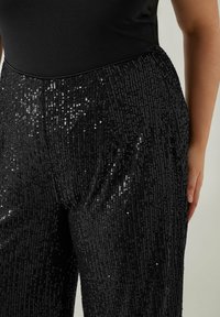 Pantalon noir pailleté avec une taille lisse, présentant des motifs verticaux de paillettes réfléchissantes et une coupe droite.