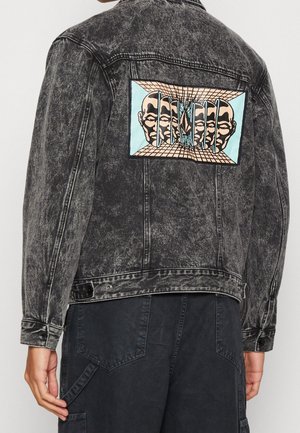 Veste en jean noire acid-wash avec un grand patch coloré dans le dos montrant plusieurs visages humains séparés par des lignes verticales dans un espace en forme de grille.