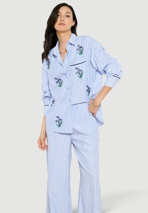 Kobieta ubrana w luźną, jasno-niebieską koszulę w paski oraz dopasowane spodnie z małymi haftowanymi kwiatowymi wzorami, pozująca z jedną ręką w kieszeni.