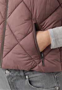 Main dans la poche d'une veste matelassée marron avec une fermeture éclair visible, portée sur un haut à manches longues gris clair et un jean gris.
