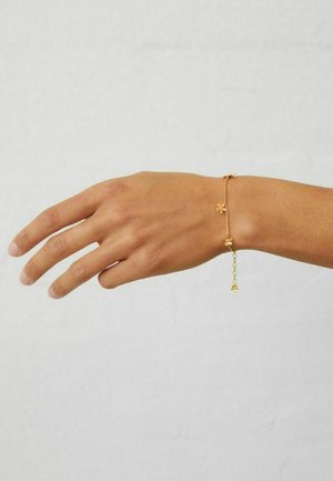 Forearm with a delicate gold chain bracelet featuring small flower-shaped charms, against a neutral background.