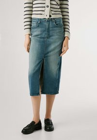 Denim kokerrok in vervaagd blauw, met een split aan de voorkant, middenlange snit en een klassieke taille. Gecombineerd met zwarte loafers.