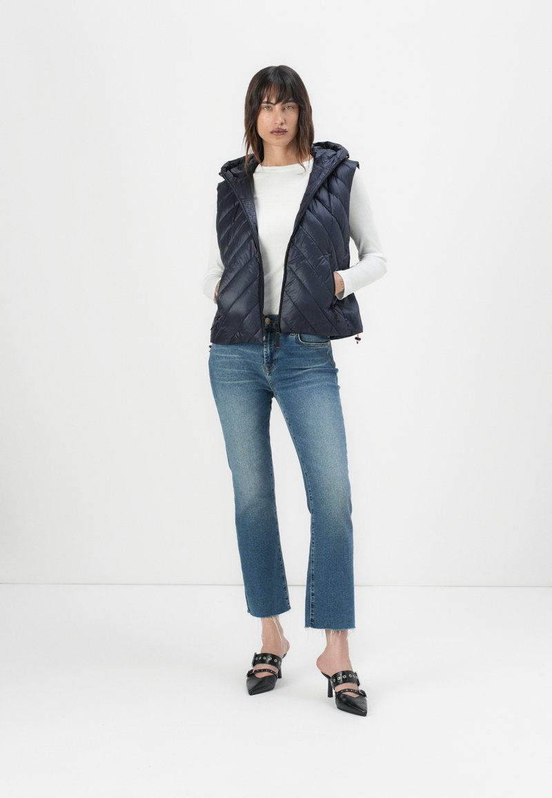 Marineblauer, gesteppter Pufferwesten mit Kapuze, getragen über einem weißen Langarmshirt. Kombiniert mit hellblauen, verkürzten Jeans und schwarzen, hochhackigen Mules.