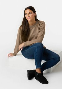 Lichte gebreide trui met geribbelde manchetten, blauwe skinny jeans en zwarte enkellaarsjes met zijaccenten, zittend op een wit platform.