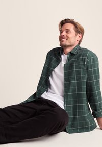 Green checkered shirt made of soft fabric, featuring gray lines, button-down collar, and a chest pocket, worn over a white t-shirt and black pants.