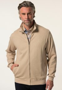 Giacca beige con zip, in tessuto texturizzato, colletto rialzato, due tasche frontali e un logo discreto sul petto.