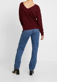 Burgunderfarbener Strickpullover mit einem V-förmigen Rücken, kombiniert mit blauen Jeans. Schwarze, hochhackige Schuhe runden das Outfit ab.
