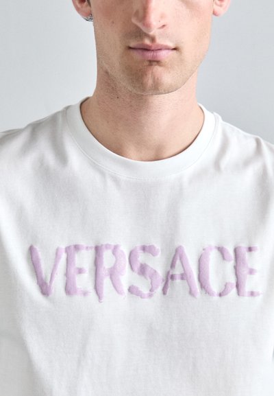 Balta medvilninė marškinė su ryškiu rožiniu-purpuriniu iškilniu "VERSACE" logotipu. Apvalus kaklas, trumpomis rankovėmis, lygus paviršius.