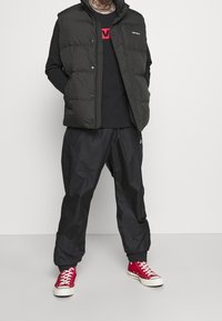 Svart pufferväst med rutigt mönster, bärs över en svart långärmad skjorta. Matchad med svarta byxor och röda sneakers.