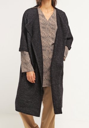 Femme portant un cardigan long gris foncé ouvert sur un chemisier beige à motifs et un pantalon marron clair, debout devant un fond uni.