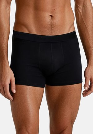 Mannelijke torso met aansluitende zwarte boxershorts, staand met ontspannen armen langs het lichaam tegen een effen achtergrond.