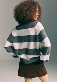 Donna con capelli ricci, che indossa un maglione a righe larghe grigio e bianco e una gonna in velluto a coste marrone, in piedi con le mani sui fianchi, di lato.