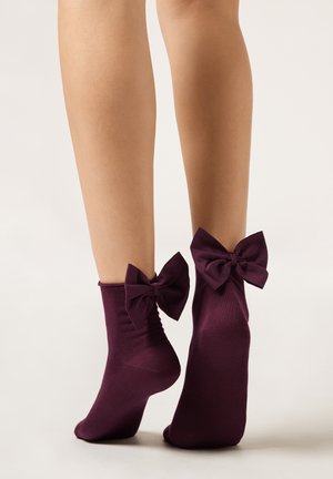 Jambes portant des chaussettes violettes aux chevilles, ornées de grands nœuds en tissu assortis à l'arrière, se tenant sur la pointe des pieds sur un fond uni.