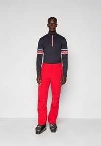 Fekete hosszú ujjú felső fehér és piros csíkokkal; piros sí nadrág; fekete csizma. Karcsúsított fazon cipzáras gallérral és zsebekkel a nadrágon.