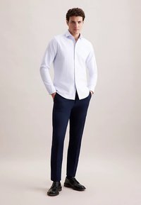 Witte, lange mouwen blouse met subtiele textuur, gedragen met donkerblauwe, slimfit broek en zwarte schoenen, handen in de zakken.