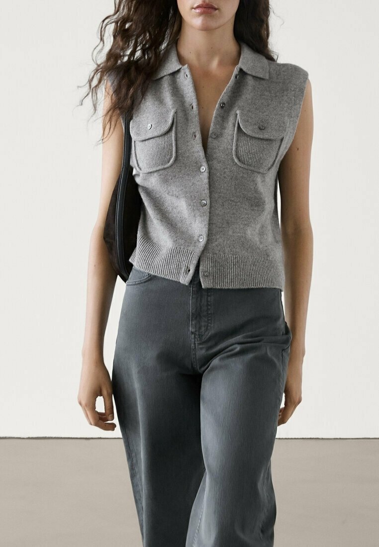 Femme portant un top en maille gris sans manches à boutons avec deux poches poitrine et un pantalon gris foncé taille haute, tenant un sac noir.