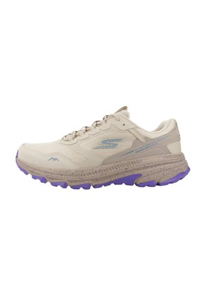 Zapatilla de running de color beige claro con parte superior de malla, que presenta acentos morados en la suela y el talón. Detalles texturizados notables y una puntera reforzada.
