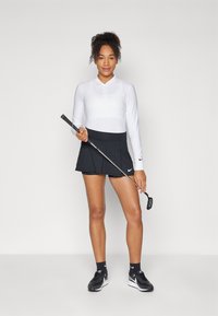 Bílá dlouhá rukávová polo košile s knoflíkovou légou, černá sukně na golf a golfová hůl. Černé tenisky a ponožky dotvářejí vzhled. Jednoduchý a sportovní design.
