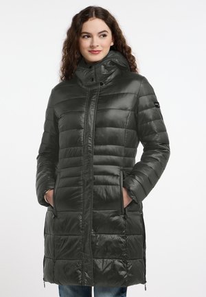 Frau mit lockigem Haar, die einen langen dunkelgrünen gesteppter Wintermantel mit hohem Kragen trägt und die Hände in den Taschen hat.