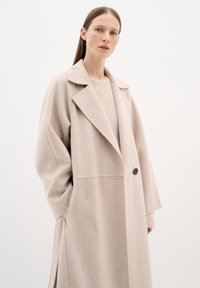 Cappotto in misto lana beige con colletto a punte, vestibilità morbida e tasche laterali. Presenta un singolo bottone nero e una texture morbida.