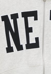 Gros plan sur un tissu gris clair avec de grandes lettres noires "N" et "E" cousues, à côté d'une fermeture éclair argentée.