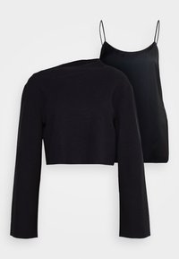 Czarny krótki sweter z długim rękawem, wykonany z dzianiny, zestawiony z czarną satynową sukienką na cienkich ramiączkach na jednolitym tle.