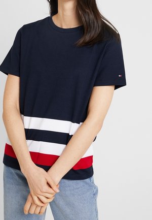 Nainen, joka pukeutunut tummansiniselle T-paidalle, jossa on valkoiset ja punaiset vaakaraidat alaosassa, yhdistetty vaaleansinisihin farkkuihin, kädet ristissä edessä.