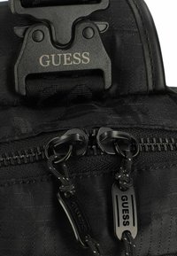 Borsa in tessuto nero con finitura testurizzata, caratterizzata da una prominente fibbia in plastica nera con la scritta "GUESS" e doppi cursori neri per la zip.