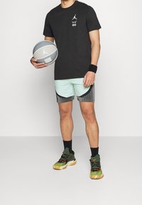Svart t-shirt med logotyp, mintgröna och grå shorts med detaljer, samt gröna och orange sneakers. Håller en grå basketboll.