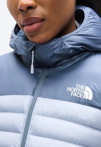 Μπλε μπουφάν με κουκούλα, με αντίθετες ανοιχτό μπλε πινελιές, με υφή καπιτονέ και λευκή λεπτομέρεια με λογότυπο. Φερμουάρ με γαντζάκι.