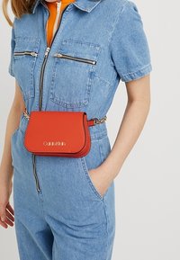 Sac en cuir rouge à la forme incurvée, avec une bandoulière chaîne et un logo embossé. Porte à la taille par-dessus une combinaison en denim. Détails de fermeture éclair sur la combinaison.