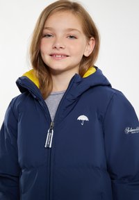 Giacca puffy blu navy con cappuccio, caratterizzata da un rivestimento giallo, logo dell'ombrello argentato e chiusura con zip. Texture liscia e design moderno.