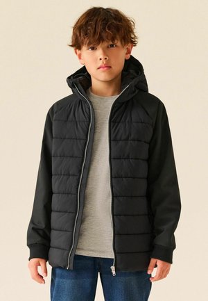Dreng med brunt hår iført en sort quiltet jakke med hætte over en grå skjorte og blå jeans, stående mod en ensfarvet baggrund.